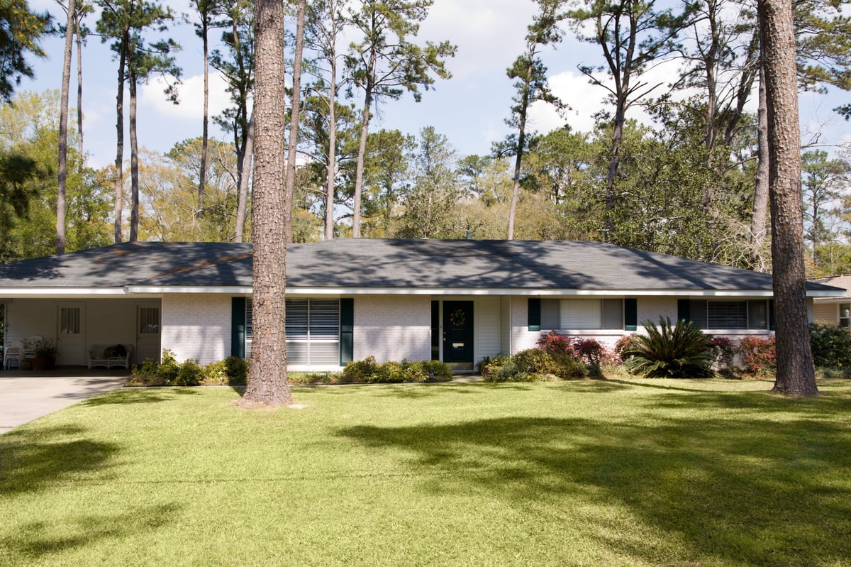Full wide-angle bright ranch-style single-family home with dark roof, surrounded by tall trees and manicured lawn on sunny day